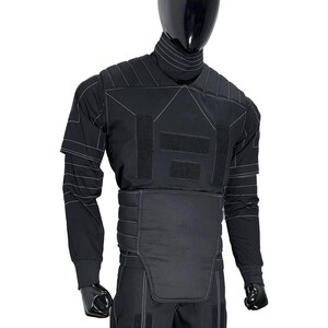 Puede incluir: Traje t&aacute;ctico negro exhibido en un maniqu&iacute;. El traje presenta un cuello alto, mangas largas y paneles reforzados con costuras blancas. El dise&ntilde;o incluye m&uacute;ltiples cierres de velcro y una secci&oacute;n media acolchada.