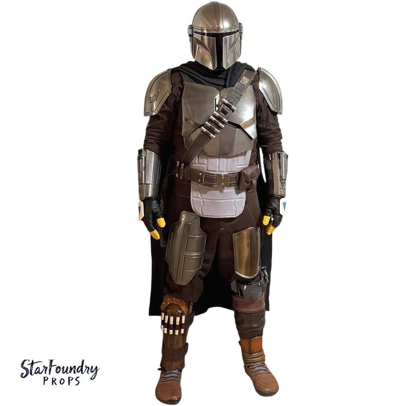 Puede incluir: Un disfraz de cuerpo completo que se asemeja a la armadura mandaloriana. El disfraz presenta un casco plateado, una armadura pectoral y hombreras. El atuendo incluye un traje interior marr&oacute;n oscuro, una capa negra y botas marrones. Incluye el texto "StarFoundry Props".