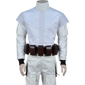 May include: A white costume featuring a high collar, long sleeves, and a brown belt with multiple pouches. The top has vertical stitching and short sleeves. The pants are white with a side pocket. Black cuffs are visible at the wrists.