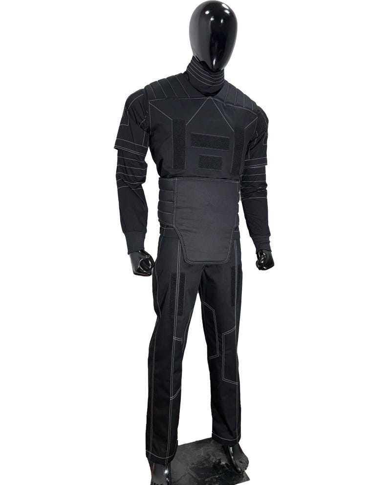 Puede incluir: Traje t&aacute;ctico negro exhibido en un maniqu&iacute;. El traje incluye una parte superior de manga larga, pantalones y un componente tipo chaleco. Las costuras blancas delinean las costuras y los paneles. El maniqu&iacute; tiene cabeza y guantes negros.