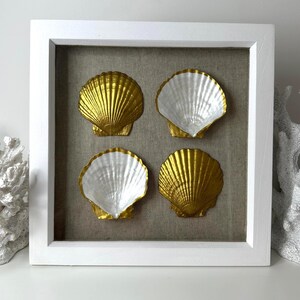 May include: A white framed artwork featuring four scallop shells. Two shells are gold, and two are white with gold edges. The shells are mounted on a neutral linen-textured background, creating a coastal theme.