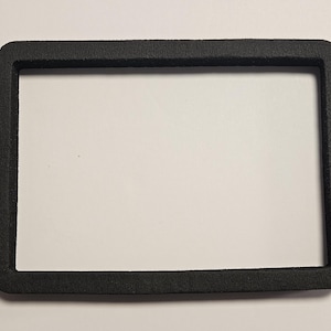 May include: A rectangular black foam frame with rounded corners and a textured surface. The object features a hollow center and a flat, uniform border design, providing a simple, open structure for various mounting or protective applications.