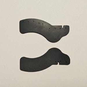 Puede incluir: Dos piezas negras y curvas, posiblemente de cuero o goma, con varios agujeros pequeños. Cada pieza tiene una forma única, una con una pequeña extensión redondeada y la otra con un diseño más angular. Sobre un fondo liso y claro.