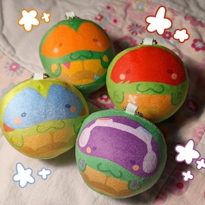 May include: Four round plush ball keychains featuring green turtle characters with different colored masks: orange, red, blue, and purple. Each plush has a small white fabric loop and a metal chain attached to the top for hanging purposes.
