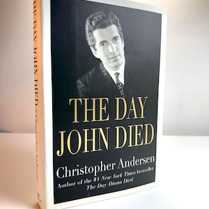 May include: A hardcover book titled "THE DAY JOHN DIED" by Christopher Andersen. The cover features a black and white portrait of a man in a suit against a black background. The title is in gold lettering.