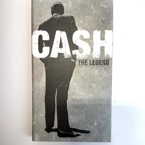May include: A book cover featuring a grayscale image of a man in a suit, viewed from the back. Bold white text reads "CASH" above "THE LEGEND". The background has a textured, distressed appearance, suggesting a vintage aesthetic.