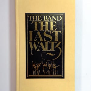 May include: A rectangular book with a pale yellow cover featuring a central black rectangular panel. The panel displays gold text reading THE BAND THE LAST WALTZ above a gold silhouette of five figures standing with their arms raised in the air.