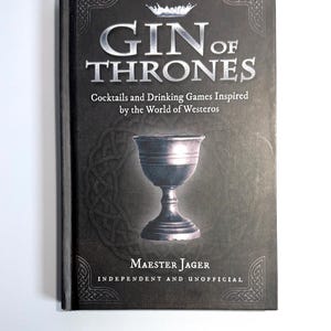 May include: A book titled "GIN of THRONES" with silver lettering and a crown graphic. The cover is dark gray with a chalice illustration and the text "Cocktails and Drinking Games Inspired by the World of Westeros." The author's name is Maester Jager.