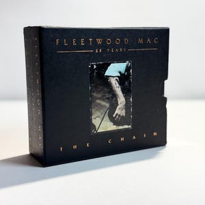 May include: Black Fleetwood Mac CD box set with gold lettering. The front features a photo of a person's arm and the text "THE CHAIN". The top of the box reads "FLEETWOOD MAC 86 YEARS".