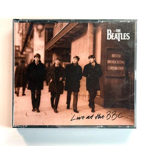 May include: A CD case featuring The Beatles Live at the BBC album cover. The image shows four individuals in dark coats walking on a city sidewalk past a building with a sign that reads British Broadcasting Corporation in white text.