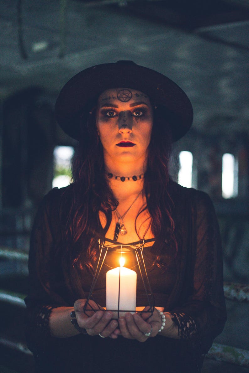 May include: A white pillar candle encased in a black geometric wire frame lantern. The candle emits a warm, bright glow, illuminating the surrounding area. The lantern features a minimalist, angular metal structure that holds the candle upright.