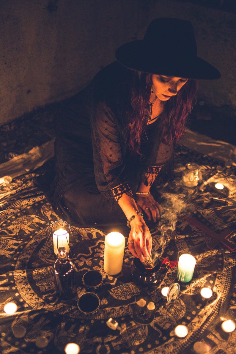 May include: A black wide-brimmed hat and a black long-sleeved top with lace trim on the cuffs. The garment is worn while holding a smoking bundle of dried herbs over a small dark bowl, surrounded by multiple lit pillar candles on a patterned cloth.