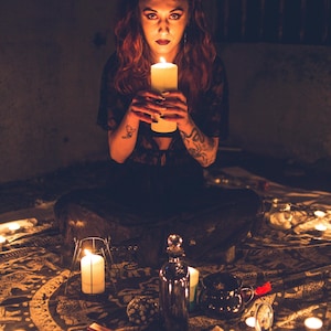 May include: A person with red hair and tattoos holding a large white pillar candle. The scene includes a dark patterned cloth, multiple lit candles, a metallic bottle, a small black cauldron, and a red cross-shaped object on the floor.