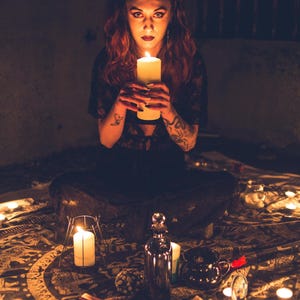 May include: A person with red hair and tattoos holding a large white pillar candle. The scene includes a dark patterned cloth, multiple lit candles, a metallic bottle, a small black cauldron, and a red cross-shaped object on the floor.