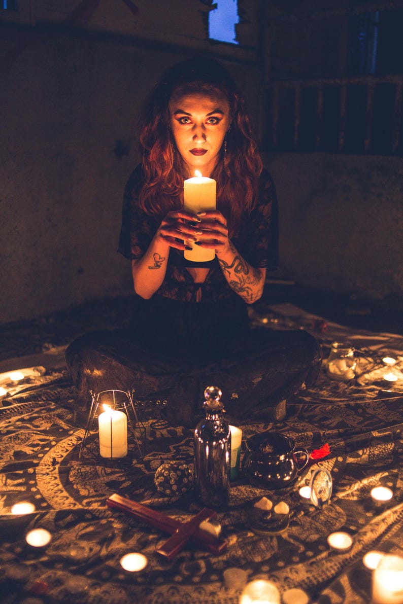 May include: A person with red hair and tattoos holding a large white pillar candle. The scene includes a dark patterned cloth, multiple lit candles, a metallic bottle, a small black cauldron, and a red cross-shaped object on the floor.