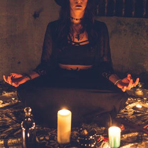 May include: A person wearing a black wide-brimmed hat, a black crop top, and a long black skirt, sitting in a meditative pose. The scene is illuminated by multiple white candles arranged on a patterned fabric, with a glass bottle and small objects.