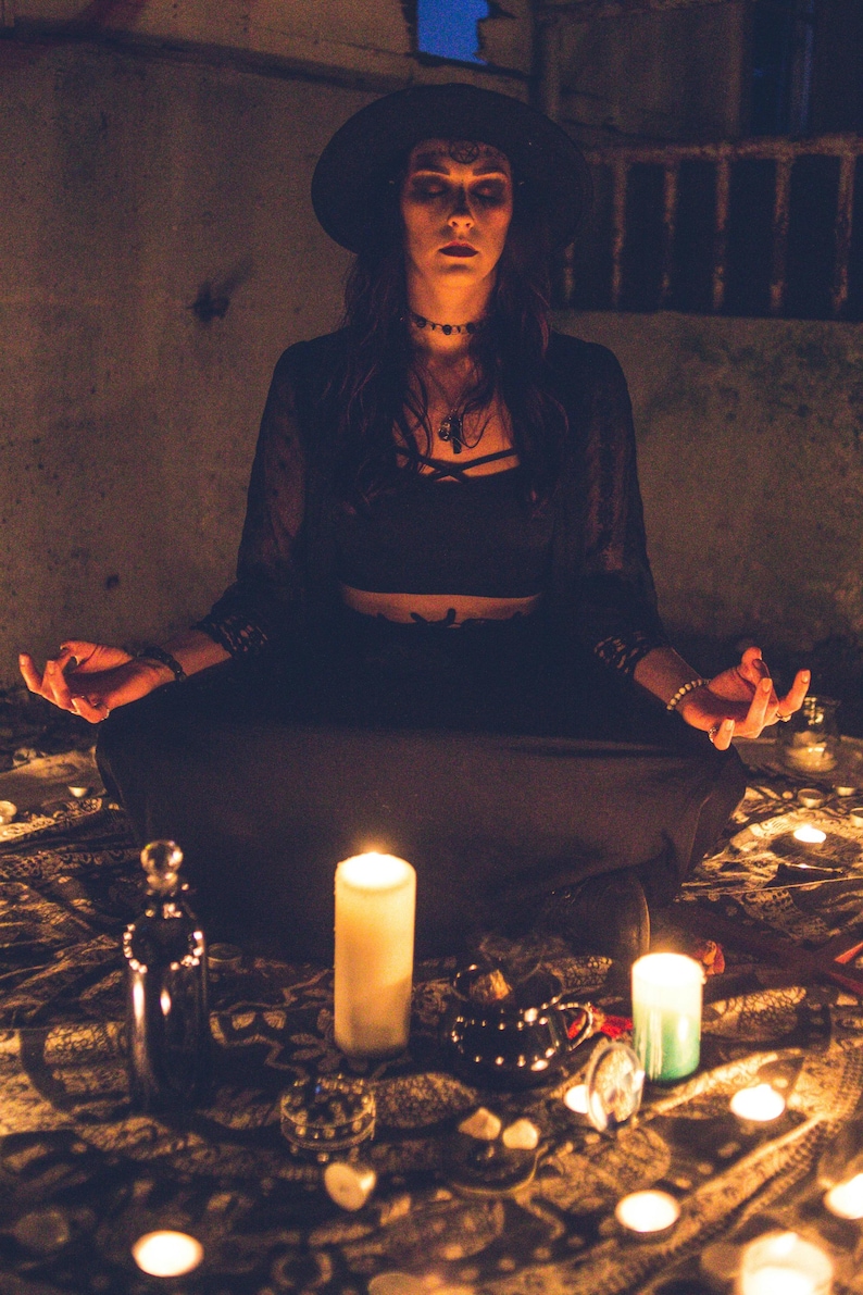 May include: A person wearing a black wide-brimmed hat, a black crop top, and a long black skirt, sitting in a meditative pose. The scene is illuminated by multiple white candles arranged on a patterned fabric, with a glass bottle and small objects.