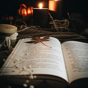 May include: An open book with a dried maple leaf resting on the pages, positioned in front of two lit black pillar candles, small decorative pumpkins, and a textured brown knit fabric, creating a warm, dimly lit atmosphere with soft candlelight.