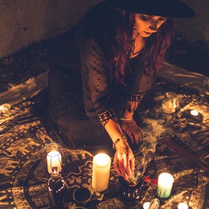 May include: A black wide-brimmed hat and a black long-sleeved top with lace trim. The garment is worn while holding a smoking sage bundle over a dark bowl, surrounded by multiple lit pillar candles on a patterned circular fabric mat.