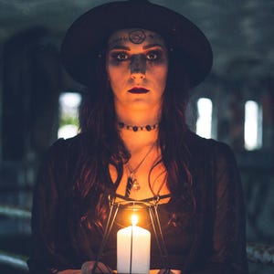 May include: A black wide-brimmed hat, a black choker, and layered necklaces worn with a black top. A white pillar candle is held inside a black geometric metal lantern frame, casting a warm orange glow on the hands and face.