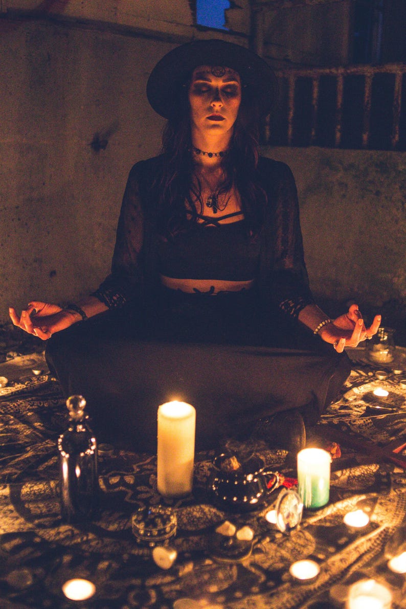 May include: A black wide-brimmed hat, a black cropped top with crisscross straps, and a long black skirt. The outfit includes a black lace cardigan, a choker necklace, and a pendant necklace, worn by a person sitting in a meditative pose.
