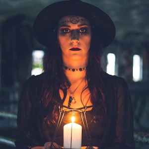 May include: A white pillar candle encased in a black geometric wire frame lantern. The candle emits a warm, bright glow, illuminating the surrounding area. The lantern features a minimalist, angular metal structure that holds the candle upright.