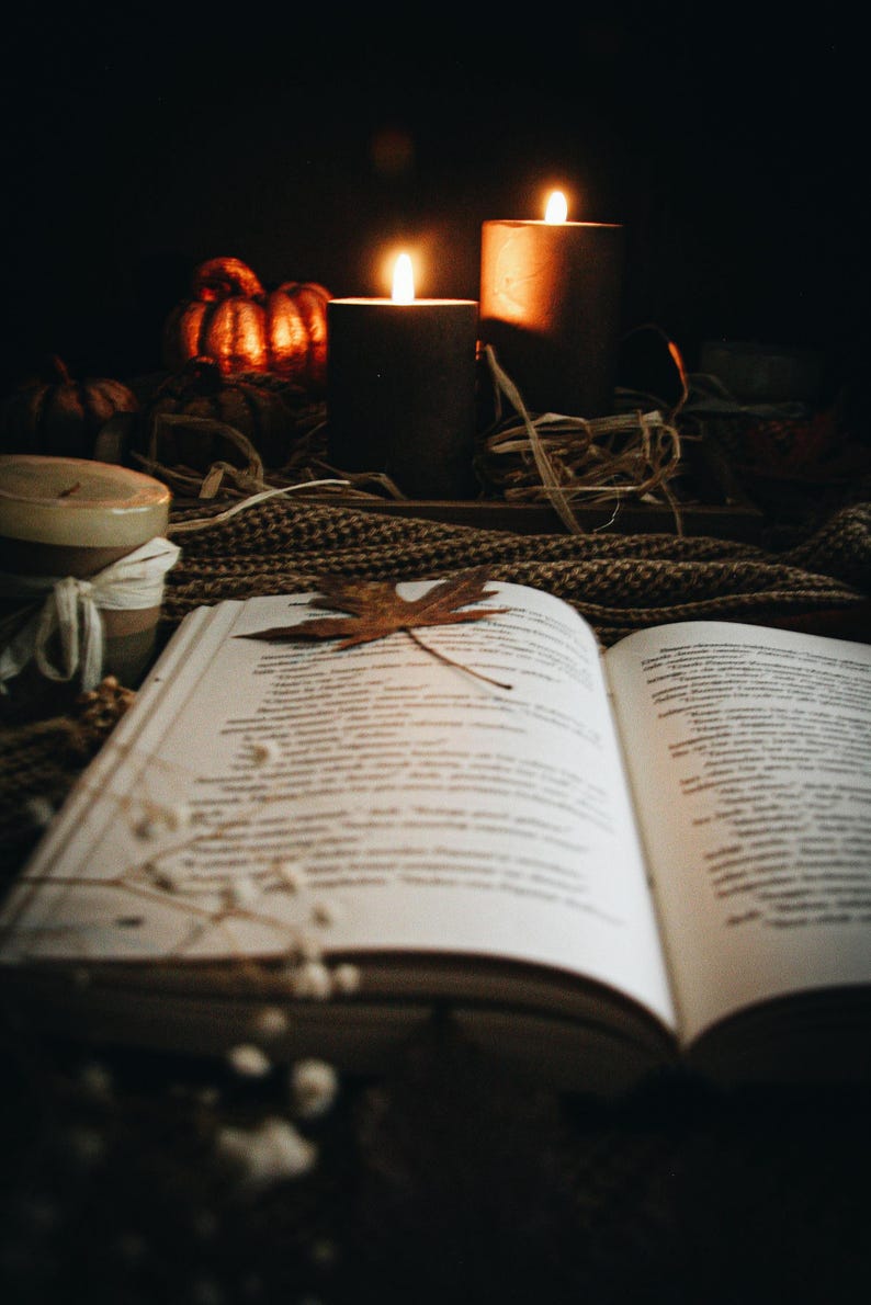May include: An open book with a dried maple leaf resting on the pages, accompanied by two lit black pillar candles, small decorative pumpkins, a brown knit textile, and a small jar wrapped in white twine, all set against a dark background.