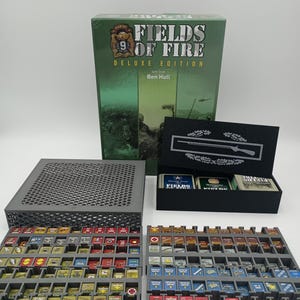 May include: A board game titled "Fields of Fire Deluxe Edition" with a green box and various game pieces. The game pieces are organized in gray trays, featuring a variety of colors and symbols. A black box with a rifle graphic is also present.