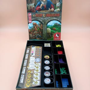 May include: A board game box set, titled "SANSA TEUTONICA BIG BOX", with a colorful illustration of a medieval scene. The box is open, revealing game components, including game boards, tokens, and wooden pieces in various colors.
