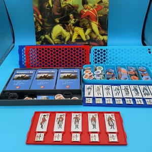 May include: A board game called "Washington's War: The American Revolution" featuring a historical battle scene on the box. The game includes strategy decks, tokens, and miniature figures. The components are stored in red and blue containers. The game is designed for strategic play.