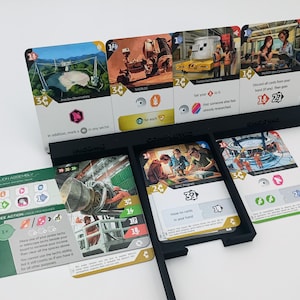 May include: A black card holder with multiple game cards. The cards feature colorful illustrations and text, with images of people, buildings, and machinery. The cards are arranged in a tiered display, with some cards standing upright and others lying flat.