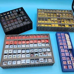 K&ouml;nnte beinhalten: Vier Spielstein-Organizer in Schwarz, Grau und Blau. Die Organizer sind mit verschiedenen Spielsteinen gef&uuml;llt, darunter quadratische Kacheln mit farbenfrohen Grafiken. Die Organizer sind so konzipiert, dass sie Spielkomponenten aufnehmen und organisieren.