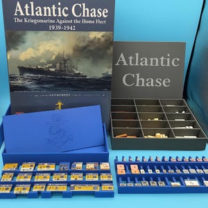 May include: A board game titled "Atlantic Chase" with a navy ship illustration on the box. The game includes a blue tray with an anchor graphic, game pieces, and a gray storage box with the game title.