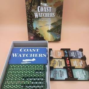 May include: A board game titled Coast Watchers featuring a box cover with a tropical scene and a person looking through binoculars. The open box contains a blue game board, a green plastic grid, and a black tray holding card decks and small pawns.