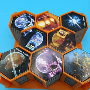 K&ouml;nnte beinhalten: Ein orangefarbener Spiel-Organizer aus Kunststoff, der Stapel sechseckiger Spielkarten h&auml;lt. Die Karten zeigen Bilder von Planeten, Weltraumph&auml;nomenen und Text wie "Plasma Storm" und "Galactic Capitol". Das Spiel scheint Weltraum-Themen zu haben.