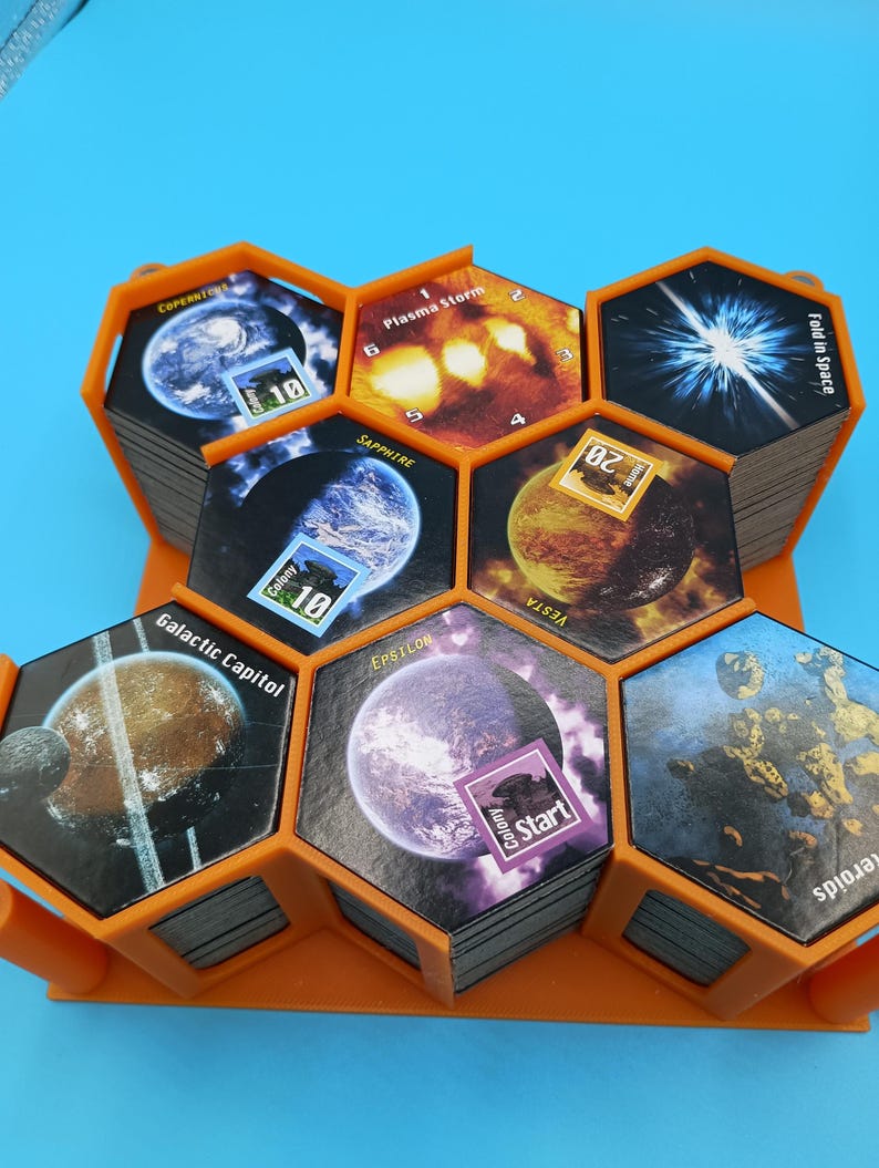 K&ouml;nnte beinhalten: Ein orangefarbener Spiel-Organizer aus Kunststoff, der Stapel sechseckiger Spielkarten h&auml;lt. Die Karten zeigen Bilder von Planeten, Weltraumph&auml;nomenen und Text wie "Plasma Storm" und "Galactic Capitol". Das Spiel scheint Weltraum-Themen zu haben.