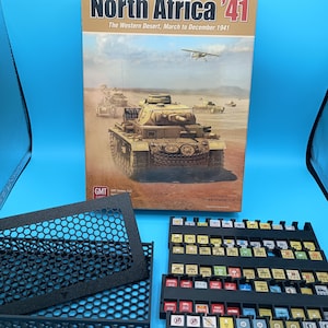 May include: A board game box titled "North Africa '41" with a desert scene featuring tanks and an airplane. The box is accompanied by black game components and a tray filled with game pieces. The game's title and subtitle are visible.