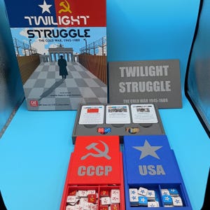 May include: A board game titled "Twilight Struggle" with a box featuring a figure walking towards a gate. The game includes cards, dice, and red and blue storage boxes labeled "CCCP" and "USA" respectively. The game's theme is the Cold War.