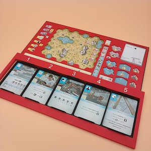 May include: A red board game with a detailed game board and a tray holding cards. The game board features a map with various spaces and icons. The tray contains cards with images and text, including "CARDS", "SPONSORS", "ANIMALS", "ASSOCIATION", and "BUILD".