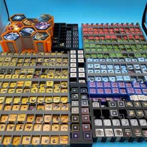 May include: A collection of board game components, including hexagonal tiles, cards, and tokens, organized in various trays. The components feature colorful graphics and text, with orange, gray, black, red, green, and blue elements. The game pieces are arranged for easy access.