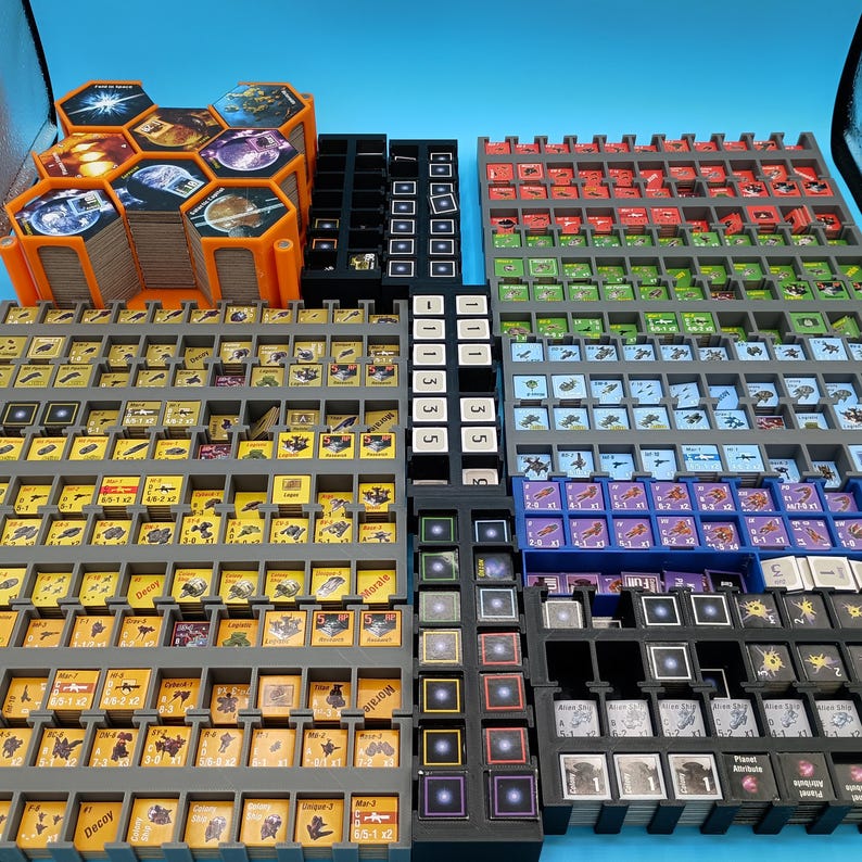 K&ouml;nnte beinhalten: Eine Sammlung von Brettspielkomponenten, darunter sechseckige Spielsteine, Karten und Spielsteine, die in verschiedenen Tabletts angeordnet sind. Die Komponenten weisen farbenfrohe Grafiken und Texte auf, mit orangefarbenen, grauen, schwarzen, roten, gr&uuml;nen und blauen Elementen. Die Spielteile sind f&uuml;r einen einfachen Zugriff angeordnet.