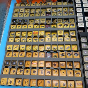 K&ouml;nnte beinhalten: Eine Sammlung von Spielsteinen, angeordnet in grauen Tabletts, mit blauem Hintergrund. Die Steine sind haupts&auml;chlich gelb und zeigen verschiedene Symbole und Texte. Die Tabletts sind in einem rasterartigen Muster angeordnet, was auf ein Brett- oder Strategiespiel hindeutet.