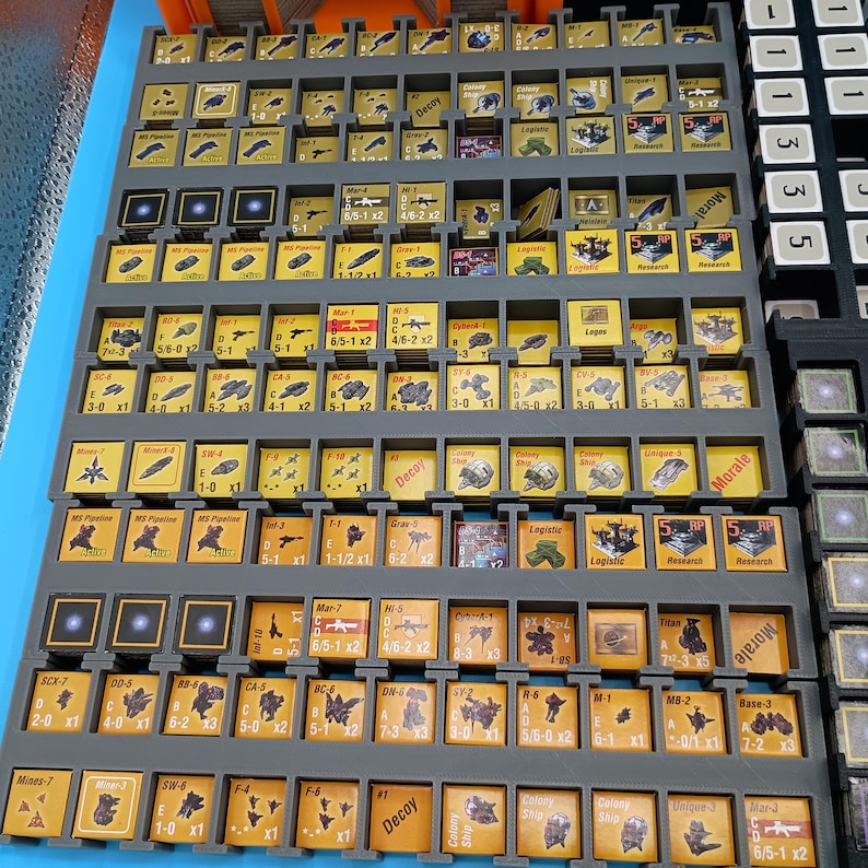 K&ouml;nnte beinhalten: Eine Sammlung von Spielsteinen, angeordnet in grauen Tabletts, mit blauem Hintergrund. Die Steine sind haupts&auml;chlich gelb und zeigen verschiedene Symbole und Texte. Die Tabletts sind in einem rasterartigen Muster angeordnet, was auf ein Brett- oder Strategiespiel hindeutet.