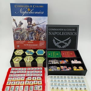 May include: A board game called "Commands & Colors Napoleonics" with a box showing soldiers. The game includes hexagonal tiles, cards, and miniature figures in red, black, and white trays. The text on the box reads "Now YOU are in command!"