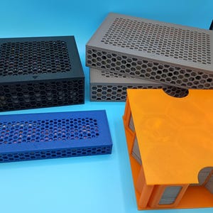 K&ouml;nnte beinhalten: Eine Auswahl an Aufbewahrungsboxen in verschiedenen Farben, darunter Schwarz, Grau, Blau und Orange. Die Boxen haben ein Wabenmuster auf den Deckeln und Seiten. Die orangefarbene Box ist offen und zeigt einen Stapel Karten.