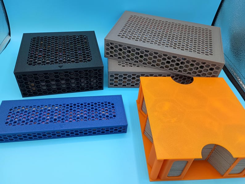 K&ouml;nnte beinhalten: Eine Auswahl an Aufbewahrungsboxen in verschiedenen Farben, darunter Schwarz, Grau, Blau und Orange. Die Boxen haben ein Wabenmuster auf den Deckeln und Seiten. Die orangefarbene Box ist offen und zeigt einen Stapel Karten.