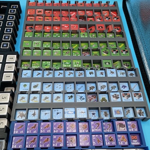 K&ouml;nnte beinhalten: Ein grauer Kunststoff-Organizer gef&uuml;llt mit kleinen, farbenfrohen Spielsteinen. Die Teile sind quadratisch und rechteckig und weisen verschiedene Symbole und Text auf. Der Organizer ist in mehrere F&auml;cher unterteilt, mit roten, gr&uuml;nen, blauen und lila Teilen.