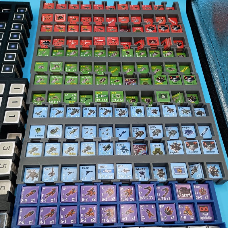 K&ouml;nnte beinhalten: Ein grauer Kunststoff-Organizer gef&uuml;llt mit kleinen, farbenfrohen Spielsteinen. Die Teile sind quadratisch und rechteckig und weisen verschiedene Symbole und Text auf. Der Organizer ist in mehrere F&auml;cher unterteilt, mit roten, gr&uuml;nen, blauen und lila Teilen.