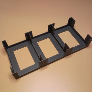 May include: A dark gray 3D-printed organizer with three rectangular openings. Each opening is framed by a raised border and supported by corner brackets. The organizer sits on a tan surface.