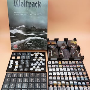 Puede incluir: Juego de mesa "Wolfpack" con una caja que presenta una escena de océano tormentoso. El juego incluye varias piezas, cartas y bandejas, todas dispuestas sobre una superficie de color naranja claro. El tema del juego son los convoyes del Atlántico Norte.